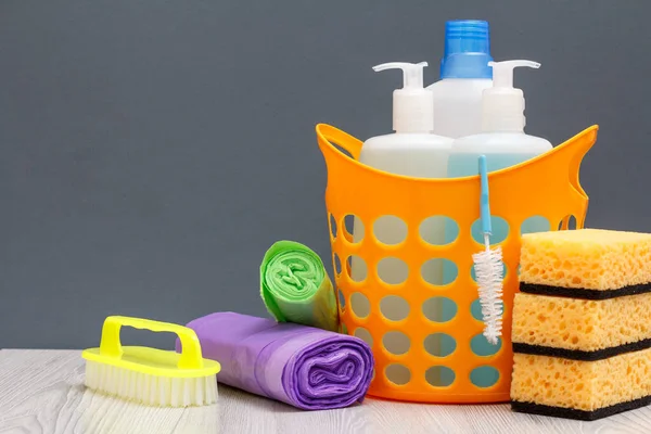 Bottles of dishwashing liquid, brush in a basket and sponges, garbage bags on gray background.