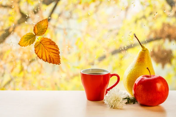 Cup of coffee, apple, pear and autumn yellow leaf on window glass with water drops in the blurred background.