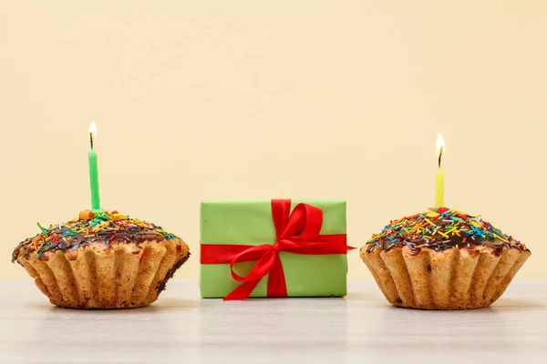 Sabrosas magdalenas de cumpleaños con velas festivas ardientes y caja de regalo sobre fondo beige . — Foto de Stock