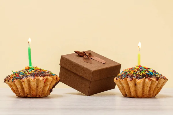 Sabrosas magdalenas de cumpleaños con velas festivas ardientes y caja de regalo sobre fondo beige . — Foto de Stock