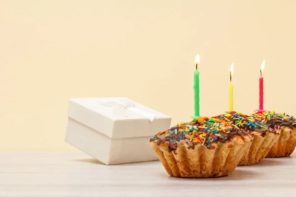 Gift box and birthday muffins with burning festive candles on beige background.