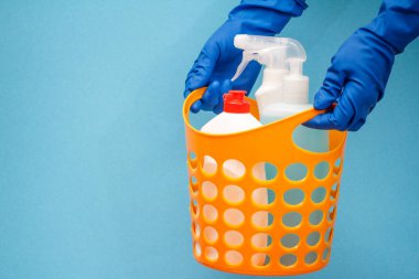 Mavi arka planda, turuncu sepette cam temizleyici şişeleri olan koruyucu eldivenli kadın elleri..