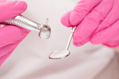 Ağız aynalı, pembe lateks eldivenli, yakın plan dişçi elleri ve arka planda yüksek hızlı diş teli. Tıbbi aletler konsepti. Sığ alan derinliği.