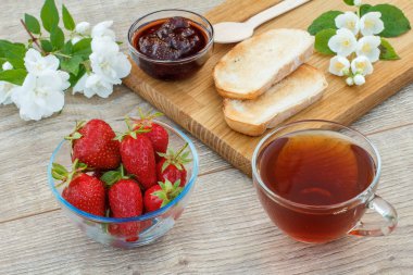 Bir bardak çay, ev yapımı çilek reçeli, ahşap kesim tahtasında ekmek ve taze çilek, kaşık ve ahşap zeminde beyaz yasemin çiçekleri. Üst görünüm.