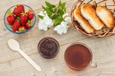 Bir bardak çay, ev yapımı çilek reçeli, taze çilek, kaşık hasır sepsiyonda kızarmış ekmek ve ahşap arka planda beyaz yasemin çiçekleri. Üst görünüm.