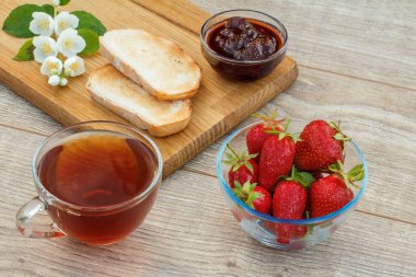 Bir bardak çay, ev yapımı çilek reçeli, ahşap kesim tahtasında ekmek ve taze çilek, ahşap zeminde beyaz yasemin çiçekleri. Üst görünüm.