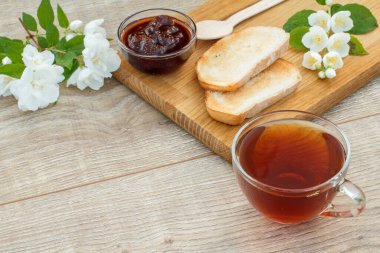 Bir bardak çay, ev yapımı çilek reçeli, ahşap kesim tahtasında ekmek ve taze çilek, kaşık ve ahşap zeminde beyaz yasemin çiçekleri. Üst görünüm.