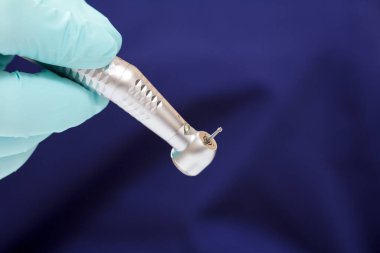 Diş hekiminin elinde yeşil lateks inceleme eldiveni ve arka planında da yüksek hızlı diş teli ve bulanık mavi tıp önlüğü var. Tıbbi araçlar konsepti.