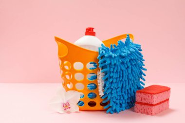 Pembe arka planda bulaşık deterjanı, bez ve süngerli plastik sepet. Yıkama ve temizleme seti.