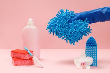 Plastik bulaşık deterjanı, sünger, fırça ve pembe arka planda bir bez parçası tutan kauçuk eldivenli bir el. Yıkama ve temizleme seti.