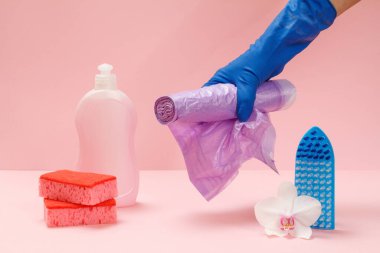 Plastik bir şişe bulaşık deterjanı, sünger, fırça ve pembe arka planda çöp torbaları tutan kauçuk bir eldiven. Yıkama ve temizleme seti.