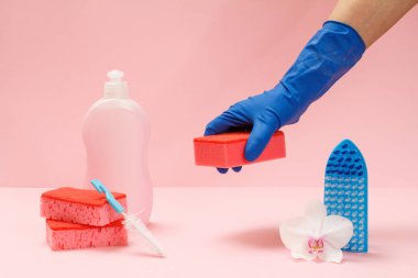 Plastik bulaşık deterjanı, süngerler, fırça ve pembe arka planda sünger tutan kauçuk eldivenli bir el. Yıkama ve temizleme seti.