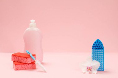 Plastik bulaşık deterjanı, sünger, fırça ve pembe arka planda bir orkide. Yıkama ve temizleme seti.
