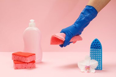 Plastik bulaşık deterjanı, sünger, fırça ve pembe arka planda bir bez parçası tutan kauçuk eldivenli bir el. Yıkama ve temizleme seti.