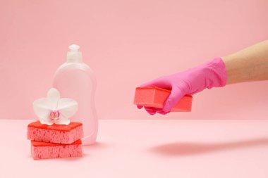 Plastik bulaşık deterjanı, süngerler ve pembe arka planda sünger tutan kauçuk eldivenli bir el. Yıkama ve temizleme seti.