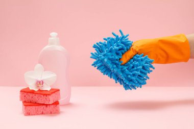 Plastik bulaşık deterjanı, süngerler ve pembe arka planda paspas tutan lastik eldivenli bir el. Yıkama ve temizleme seti.