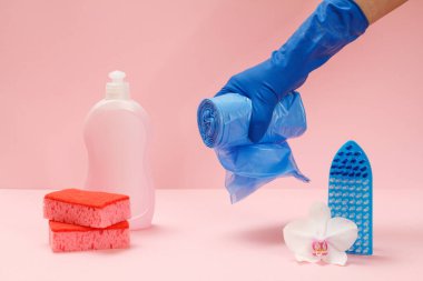 Plastik bulaşık deterjanı, sünger, fırça ve pembe arka planda mavi çöp torbaları tutan kauçuk eldivenli bir el. Yıkama ve temizleme seti.