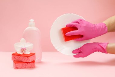 Plastik eldiven, sünger, bir şişe bulaşık deterjanı ve pembe arka planda süngerler olan kadın elleri. Yıkama ve temizlik konsepti.