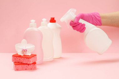 Plastik çamaşır şişeleri, süngerler ve pembe arka planda cam ve fayans temizleyicisi tutan kauçuk eldivenli bir el. Yıkama ve temizleme seti.