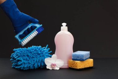 Plastik bir şişe bulaşık deterjanı, bir bez, süngerler ve siyah arka planda fırçası olan bir el. Yıkama ve temizleme seti.