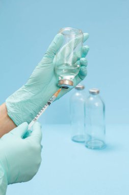 Doktorun elinde cam şişe ve şırınga tutan lateks eldivenli ellerini yakından çek ve mavi arka planda enjeksiyon için sıvı ilaç al. Sağlık ve Tıp Konsepti.