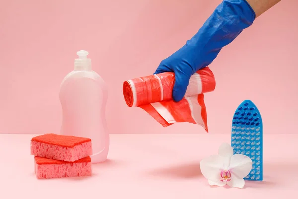 Plastic bottle of dishwashing liquid, sponges, a brush and a hand in a rubber glove holding garbage bags on the pink background. Washing and cleaning set.
