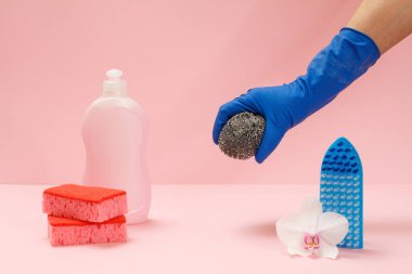 Plastik bir şişe bulaşık deterjanı, süngerler, bir fırça ve pembe arka planda metal bir sünger tutan lastik eldivenli bir el. Yıkama ve temizleme seti.