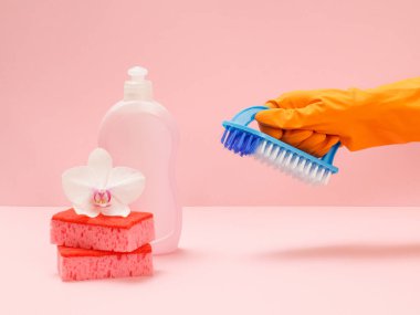 Plastik bulaşık deterjanı, sünger, beyaz bir orkide çiçeği ve pembe arka planda fırça tutan kauçuk eldivenli bir el. Yıkama ve temizleme seti.