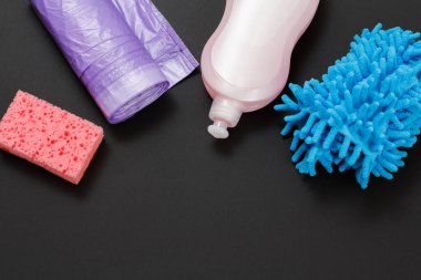 Plastik bir şişe çamaşır suyu, paçavralar, çöp torbaları ve siyah arka planda bir sünger. Üst Manzara. Yıkama ve temizleme seti.