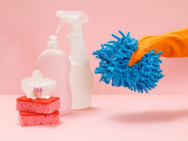 Plastik çamaşır şişeleri, süngerler ve pembe arka planda paspas tutan kauçuk eldivenli bir el. Yıkama ve temizleme seti.