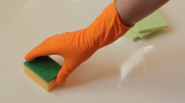 Paçavralar fayans yüzeyinde ve turuncu lastik eldivenli bir erkek eli bir sünger ve bir fırça koyuyor..