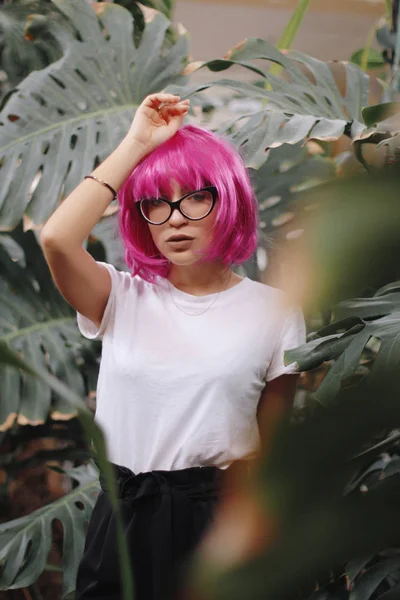 戴眼镜的女孩。丛林里的女人穿白色 t恤衫的女孩戴着刘海头粉色假发。背景植物 — 图库照片