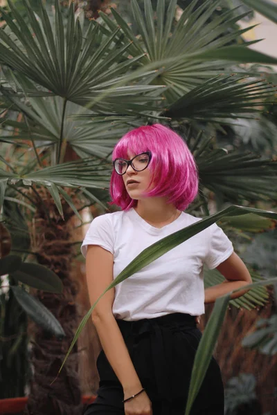 戴眼镜的女孩. 丛林中的女人穿白色 t恤衫的女孩戴着刘海头粉色假发。背景植物 — 图库照片