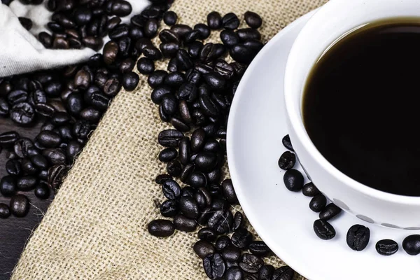 Beyaz Kupası ve kahve çekirdeği kumaş ve ahşap arka plan üzerinde siyah kahve.