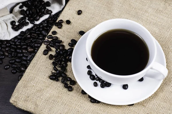 Beyaz Kupası ve kahve çekirdeği kumaş ve ahşap arka plan üzerinde siyah kahve.