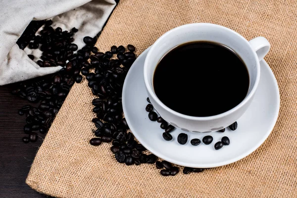 Beyaz Kupası ve kahve çekirdeği kumaş ve ahşap arka plan üzerinde siyah kahve.