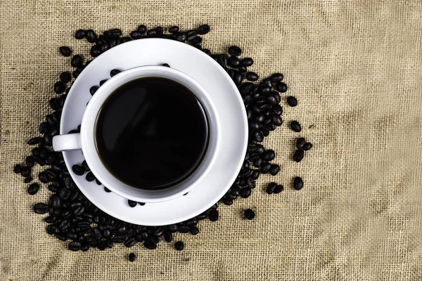 Beyaz Kupası ve kahve çekirdeği kumaş ve ahşap arka plan üzerinde siyah kahve.