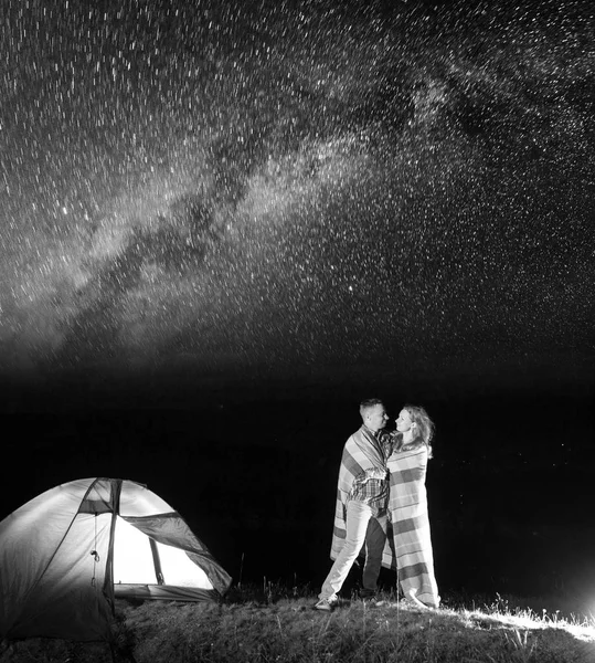 Sweety couple hikers standing together near campfire and tent at night and enjoying starry sky. Pair covered with a plaid. Amazing Milky way. Black and white