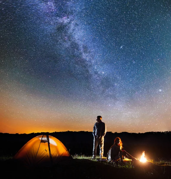 Romantický pár turistů v jeho táboře v noci u táboráku a stan proti hvězdné oblohy s mléčnou dráhu — Stock fotografie