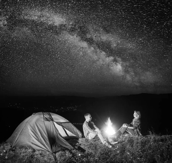 Night tent camping. Young couple hikers sitting near fire under incredibly beautiful starry sky and Milky way. In the background luminous village in the valley. Black and white — Stock Photo, Image