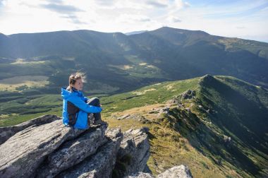 Genç bir kadın dağlarda bir kayanın üzerinde oturuyor.