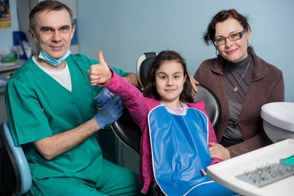 Retrato de dentista senior y niña con su madre en la visita dental en el consultorio dental. El paciente feliz sonríe y muestra los pulgares hacia arriba. Concepto de odontología, medicina y salud — Foto de Stock