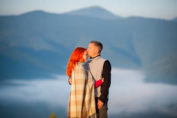 Романтична пара туристів цілує і обіймає один одного на тлі красивого гірського ландшафту з ранковим туманом над горами на фоні. Рудоволоса дівчина вкрита ковдрою. Крупним планом — стокове фото