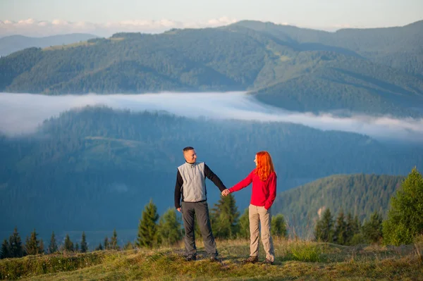 Ζευγάρι κρατώντας τα χέρια και να βλέπουν ο ένας τον άλλον να στέκεται πάνω σε ένα λόφο όμορφο φόντο με πρωινή ομίχλη πάνω από τα βουνά. — Φωτογραφία Αρχείου