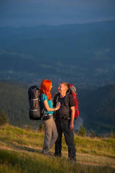 Heureux couple touristique debout sur la route dans les montagnes, se regardant et souriant sur le fond des montagnes puissantes paysagées — Photo
