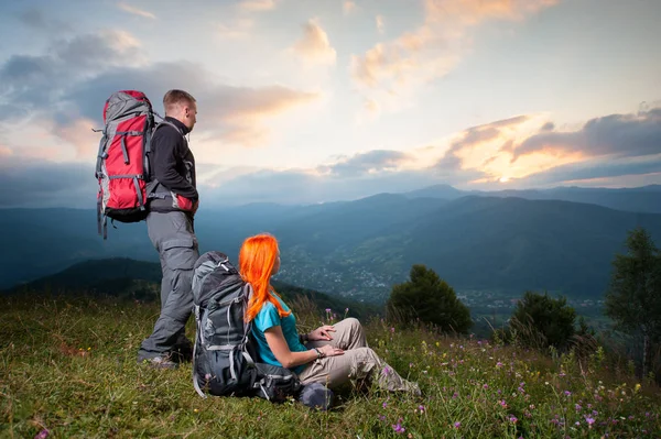 Lyckligt par vandrare med ryggsäckar som njuter av vacker utsikt på en kulle. Berg, by i dalen och molnig himmel i solnedgången på bakgrund — Stockfoto