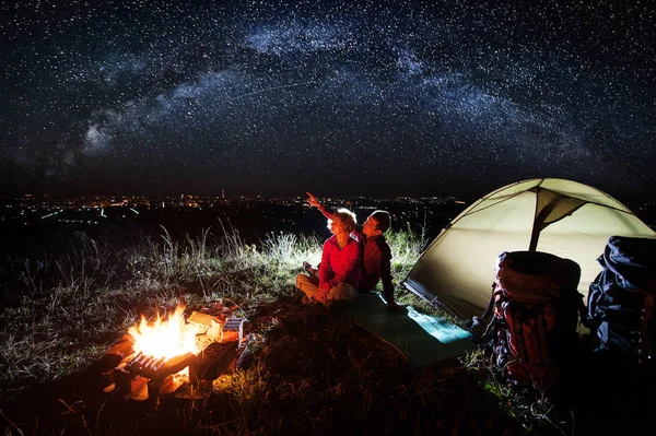 Night camping near the town. Young pair sitting near campfire and tent with backpacks, looking at beautiful night sky full of stars and enjoying night scene. Man is pointing at the sky. Long exposure