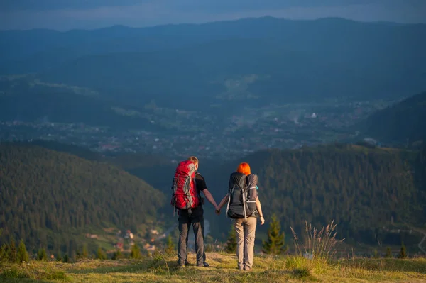 Vista posteriore coppia escursionisti con zaini che si tengono per mano, in piedi sulla cima della collina, godendo della vista di bella vista aperta sulle montagne e villaggio nella valle — Foto Stock