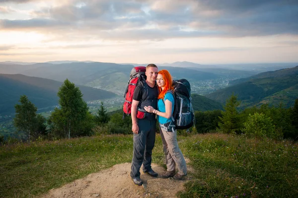 Ritratto completo di coppia turisti con zaini sta abbracciando e godendo la vista di una splendida vista sulle montagne, boschi, colline, villaggio nella valle e cielo nuvoloso — Foto Stock