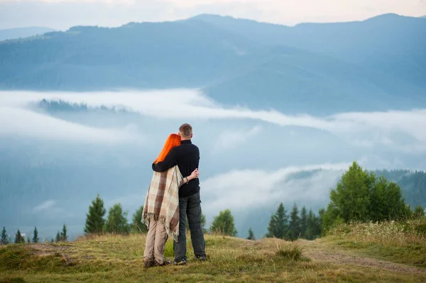 Çekici çifti zevk bir sabah bir tepede duran Dağları pus. Genç çift birbirlerine sarılma. Kızıl saçlı kız bir battaniye ile örtülü. Arkadan görünüş — Stok fotoğraf